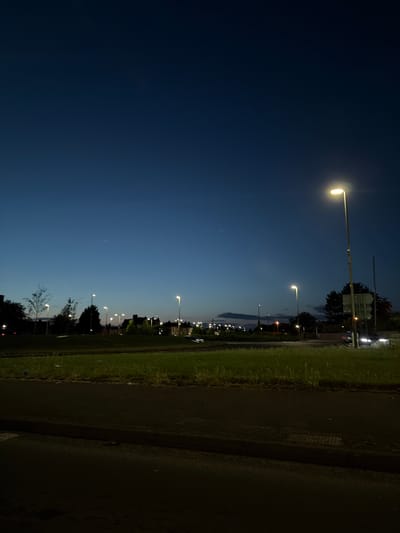 A nighttime outdoor scene, possibly a street in Leicester, UK. A row of evenly spaced streetlights illuminates a grassy verge. The lights progressively diminish in brightness into the distance, corresponding with a change in sky gradient from dark blue to a lighter, twilight hue. Trees and other vegetation are visible in silhouette against the twilight sky. A dark, paved surface is in the right foreground.