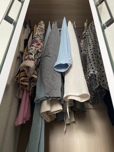 The image shows a close-up view of a shelf in a closet, packed with folded clothing items. The shelf itself appears to be made of a light-colored wood. The foreground is dominated by the edge of a white drawer or shelf front, with reflective glass on top.
The clothing is stacked horizontally and vertically. At the top, a grey fabric with a black polka dot pattern is visible, draped over other items. Below this, there's a neatly folded white textured garment, followed by a light blue garment with a button detail. A grey knitted sweater is stacked on top of a light blue fabric item. Further down, a stack of folded clothes includes a garment with a floral and bird print, a striped item, and some solid-colored fabrics in pink and yellow. A fuzzy white textile is visible at the bottom of the frame.
The lighting suggests an indoor setting, possibly during the daytime. There is no visible text in the image. The overall impression is one of organized storage of textiles. The scene is set within a domestic environment, likely a bedroom or dressing area. The clutter suggests recent use or preparation for storage.