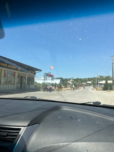 This is a photograph taken from inside a vehicle, looking out through the windshield. The image is oriented vertically, with the sky taking up the left two-thirds and the road and buildings on the right.
The sky is a clear, bright blue, suggesting a sunny day. There are some wispy white clouds scattered across it. The view from the car shows a rural or semi-urban setting. A paved road is visible, with some unpaved areas and gravel on the sides. Along the road, there are various structures, including buildings with visible signs and what appears to be a construction site with scaffolding. Trees and greenery are scattered throughout the scene.
In the distance, a tall structure with scaffolding is visible, topped with a red and orange flag. Further down the road, a red motorcycle is parked or being ridden by someone. There are also a few figures on bicycles in the middle ground, suggesting ongoing activity. The building closest to the foreground has a large sign with text that appears to be in a Cyrillic script. Parts of the text are legible: "ТУРПИК ДУКАНЫ" which translates to "Fruit Store" or "Produce Store".
The time of day appears to be midday, given the bright sunlight and the shadows cast. The overall impression is one of a typical day in a populated area, with people going about their business. The image seems to capture a moment during travel, with the dashboard of the car partially visible in the foreground.
