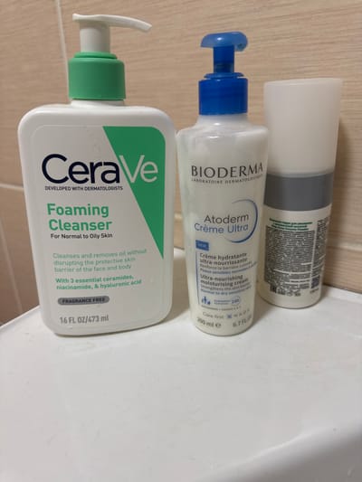 Three cosmetic product bottles are arranged on a white surface, which exhibits minor liquid residue, against a light, horizontally textured background wall. From left to right, the objects are: a white CeraVe Foaming Cleanser pump bottle with a green pump collar, indicating a volume of 16 FL OZ / 473 mL; a white BIODERMA Atoderm Crème Ultra pump bottle with a blue pump top, specifying a volume of 200 ml e / 6.7 FL.OZ; and a partially visible cylindrical bottle with a grey lower section and a white opaque upper section, displaying Cyrillic script on its label. No people, actions, or interactions are depicted. The scene is located in Ciorescu, Moldova.