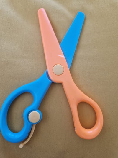 The image shows a pair of orange and blue plastic safety scissors lying on a tan, textured surface, possibly fabric. The scissors are open, with the blades pointing upwards. The left handle is blue, and the right handle is orange. There is a light-colored pivot point where the blades and handles meet. The background is out of focus. The image's origin is in Fresnes, France.
