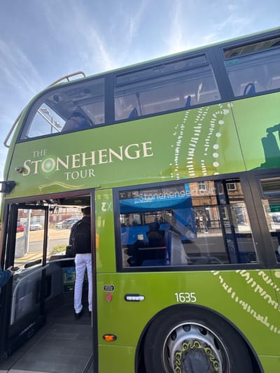 A lime green double-decker bus, identified as "THE STONEHENGE TOUR" with fleet number 1635, is depicted from a low, upward-angled perspective on a street in Salisbury, United Kingdom. The bus is stationary with its front passenger door open. A male individual, wearing a black backpack, dark jacket, white trousers, and black shoes, is in the process of boarding the bus, seen from behind. The bus features large panoramic windows on both its upper and lower decks. The upper deck interior shows an obscured passenger. The lower deck windows reflect the opposite side of the street, revealing brick buildings, a pavement, several pedestrians, and a second bus, which is blue and also appears to be a "Stonehenge Tour" vehicle. Through the open bus door, a background street scene with parked cars (including red and grey vehicles) and low-rise structures is visible. The sky is blue with scattered wispy clouds, indicating daylight conditions.