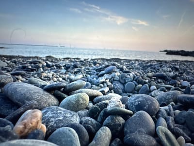The image shows a close-up view of a rocky beach. The foreground is dominated by smooth, varied-sized stones in shades of gray, blue, and some with lighter speckles. The stones are densely packed, creating a textured surface. In the background, the sea meets the horizon under a pale sky that transitions from a soft pinkish hue near the horizon to a muted blue overhead. Wispy clouds are visible in the sky. The overall scene suggests a calm, natural coastal environment. This image does not depict the city of Porrentruy, Switzerland, as Porrentruy is an inland town and not a coastal location.