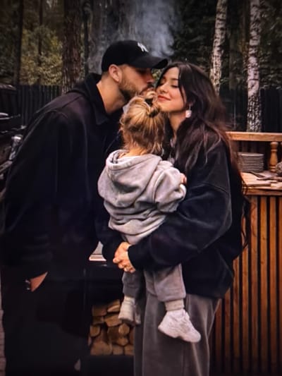 The image captures a tender moment between a couple and a young child. The scene appears to be outdoors, with a rustic wooden structure in the background, possibly a cabin or porch. The subjects are positioned closely, with the woman embracing the child, who is nestled between her and the man. The man is kissing the woman, and the child is leaning in, their faces close.

The woman has long, dark hair and is wearing a dark sweatshirt and grey sweatpants. She has a gentle smile and her eyes are closed. The child has lighter hair styled in a bun and is also wearing a grey hoodie. The man is wearing a black baseball cap and a black sweatshirt, and has a beard. There is a faint wisp of smoke or steam visible near the man's head.

The lighting suggests it might be dusk or a dimly lit day. The overall mood is intimate and affectionate, conveying a sense of family warmth and connection. The background shows a blurred natural environment with trees and a wooden wall with horizontal slats, which may be part of the rustic structure. The ground appears to be covered in a dark material, possibly soil or wood chips. There is no visible text in the image.