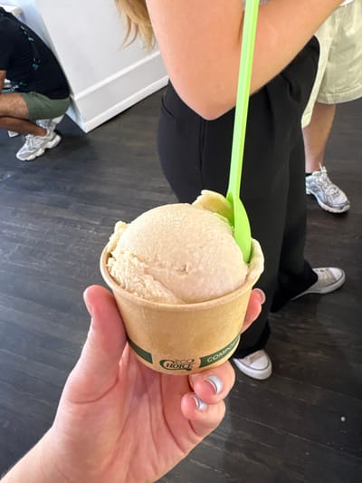 A light-skinned hand, with fingernails painted in a light blue French manicure style, holds a beige paper cup containing a scoop of light beige ice cream or gelato. A bright neon green plastic spoon is inserted into the ice cream. The cup features green text, including "ECO CHOICE" and "COMPOSTABLE," and a green stripe around its lower circumference.
Behind the hand and ice cream, the upper arm and lower torso of a person wearing black pants are visible, with white low-top sneakers on their feet. To the left, a person wearing a dark t-shirt and green shorts is in a squatting position against a white wall with a gray baseboard, wearing white athletic shoes. The lower legs and feet of another person, wearing light shorts and gray athletic shoes, are visible further in the background to the right. The floor is dark brown, appearing to be wood or laminate. The scene is located indoors in Lambertville, United States.