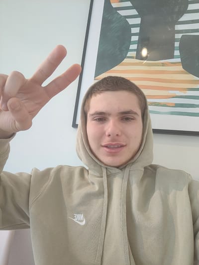 A young man wearing a tan hooded sweatshirt with the word "NIKE" stitched in white on the left breast is making a peace sign with his left hand. He appears to be indoors, with a white wall behind him and a framed piece of artwork hanging to his right. The artwork features a pattern of horizontal stripes, with some dark green and black shapes overlaid. The location in the city of Waregem, Belgium, is not evident from the image.