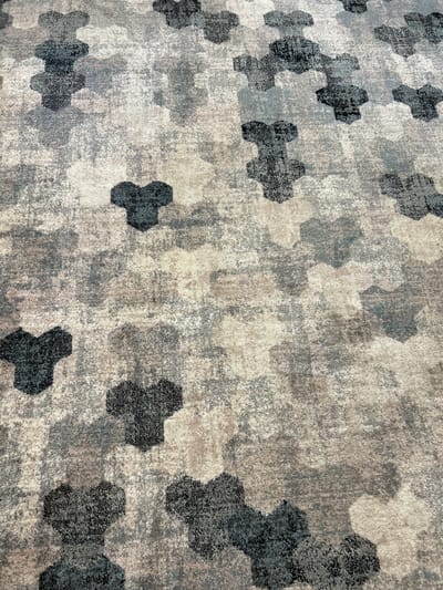 The image shows a close-up, elevated view of a textured carpet with a repeating hexagonal pattern in shades of grey, black, and beige. The hexagons appear as if they are layered or overlapping, creating a sense of depth and visual interest. The texture of the carpet is slightly rough, with individual fibers visible, suggesting it is a woven material. The lighting casts subtle shadows, highlighting the raised areas of the pattern and adding to the overall depth. There are no people, animals, or any recognizable objects other than the carpet itself. The scene appears to be indoors, likely a floor covering in a domestic or commercial space. There is no specific identifiable location within the city of Pavlohrad, Ukraine, visible in this image.