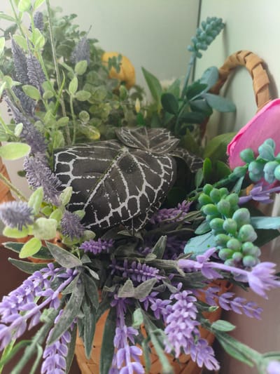 The image shows a close-up view of a basket filled with artificial flowers and greenery. The flowers include purple lavender-like blooms and clusters of small green buds. There are also some grey-green leaves with a striking white veined pattern, and a hint of a pink petal. The basket itself appears to be made of woven material, with a textured handle visible at the top right. The background is a plain, light-colored surface, possibly a wall. The lighting is soft, casting a slight shadow on the background. There are no people or animals present in the image. The context of "Азьмушкино, Russia" is not visually supported by the image content.