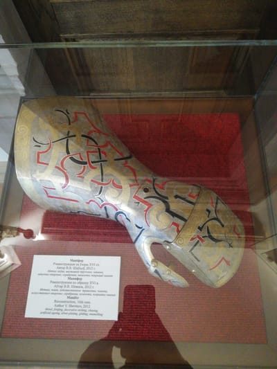 A reconstructed 16th-century "Manifer" (gauntlet) is displayed within a glass exhibition case in a museum setting at the Mir Castle Complex, Мірскі сельскі Савет, Belarus. The metal gauntlet, crafted by V.V. Shevtsov in 2012 using forging, decorative etching, chasing, artificial aging, silver-plating, gilding, and enamelling, features intricate red and black enameled patterns over a gilded and silver-plated surface with etched spiral motifs. It is positioned at an angle, resting on a textured reddish-brown fabric background. A white rectangular information label in Belarusian, Russian, and English, detailing the artifact's reconstruction, author, and materials, is placed in front of the gauntlet. A faint, distorted reflection of a person's upper body is visible on the lower right glass panel of the display case. The background above the exhibit appears to be dark, possibly wooden.