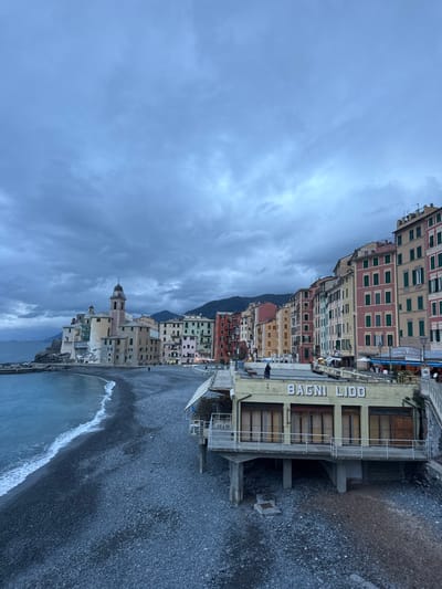 The image depicts the coastal town of Camogli, Italy, under an overcast sky. In the foreground, a dark pebble beach extends across the lower portion of the frame, with gentle waves breaking along the shoreline on the left. A two-story building, identified by the large white letters "BAGNI LIDO" on its facade, stands partially over the beach on concrete stilts, featuring multiple closed window/door panels on its ground level. A single individual, dressed in dark clothing, stands on the rooftop terrace of the "BAGNI LIDO" building, looking towards the right. An elevated walkway with stairs connects to the right side of this structure.

In the midground, a prominent row of tall, narrow, multi-colored buildings, ranging from pinks, reds, and yellows to greens and creams, lines the waterfront, ascending the hillside from left to right. Many windows feature dark green shutters. To the far left, built on a rocky promontory, stands a large, light-colored church with a domed roof and a clock tower, flanked by other pale-colored structures. A short breakwater extends into the sea from the church's base. Several faint lights are visible from businesses at the base of the colorful buildings. Two individuals are discernable on the pebble beach; one is walking near the waterline on the left.

The background features dark, forested hills rising behind the town. The sky is uniformly covered with thick, grey-blue clouds, indicative of overcast conditions or dusk. The sea, a deep blue, meets the cloudy horizon on the far left.