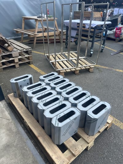 The image depicts an outdoor industrial or workshop setting, likely in Loo, Estonia. In the foreground, a pallet holds a collection of identical, hexagonal, silver metal objects with cut-out openings. These objects are arranged in a grid-like pattern. Behind them, a larger metal rack structure stands on wooden pallets. To the left, a stack of wooden pallets is visible, partially obscuring a grey tarp or wall. In the background, a workbench with a wooden top and metal legs is present, along with other industrial equipment and materials. The ground is paved with asphalt, marked with yellow lines. There are no people visible in the image.