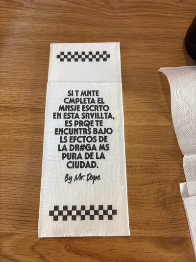 The media file is a close-up, top-down shot of a white fabric tag with black text and checkerboard patterns. The tag is lying on a wooden surface, likely a table. The text on the tag appears to be in Spanish, with a poem or message about a "city" and its "effects." The message includes phrases like "SI T MNTE CMPLETA EL MNSJE ESCRTO EN ESTA SRVILLTA" which translates to "IF THE COMPLETE MESSAGE WRITTEN IN THIS VILLAGE," and "LS EFCTOS DE LA DR#GA MS PURA DE LA CIUDAD" which translates to "THE EFFECTS OF THE PUREST DRUG OF THE CITY." The tag also features the signature "By Mr. Dope." The checkerboard patterns are on the left and right edges of the tag, suggesting a brand or design element. The overall impression is of a garment tag or a similar piece of fabric branding. The wooden surface suggests an indoor setting, possibly a home or a shop. The lighting is even, indicating it might be daytime or under artificial light.