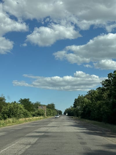 The image captures a road scene in Hîrbovăț, Moldova, under a partly cloudy sky. The perspective is from a moving vehicle, looking forward down a two-lane asphalt road. The road surface shows signs of wear, with patches and tire marks visible. Lush green trees and foliage line both sides of the road, with taller trees forming a dense canopy on the right and a mix of trees and grass on the left. In the distance, a white car is traveling on the road. On the left side of the road, there's a billboard with some graffiti or writing on it. The overall impression is a rural or semi-rural setting during the daytime.