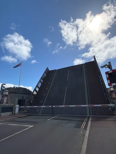 The image shows the raised Weymouth Town Bridge in Weymouth, United Kingdom, viewed from a low angle. The blue sky with scattered white clouds fills the background. The bridge is a bascule bridge, with the road surface lifted at an angle, revealing the underside. The bridge barriers are down, preventing road traffic. A Union Jack flag flies from a pole attached to a nearby building. The sun is high in the sky, with lens flare present.