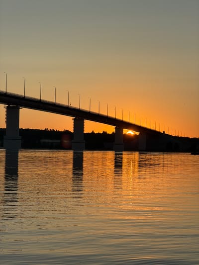 This is a vertical, medium shot taken from a body of water, likely the Kama River, looking towards a long bridge at sunset in Kambarka, Russia. The sky is a gradient of soft orange and pale blue. The bridge spans the width of the frame, with its pylons casting reflections on the water. The sun is partially obscured by the bridge, creating a bright orange glow and a shimmering reflection on the water. A dark treeline is visible in the distance behind the bridge, silhouetted against the sunset. The water is calm, with gentle ripples. There are no people visible in the image.