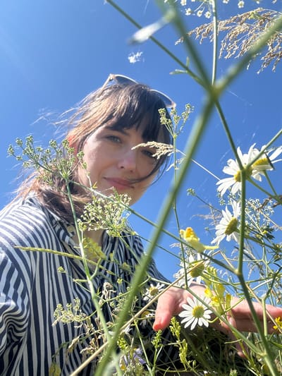 The image shows a person from a low angle, looking up at the sky and through wildflowers. The person has dark hair and bangs, and is wearing sunglasses on their head and a striped shirt. They are reaching out with their right hand towards the flowers. The background is a clear blue sky, with some wispy clouds and tall grasses. The scene appears to be outdoors, in a field or meadow. There is no specific indication of the location being in Krasnyi Yar, Russia.