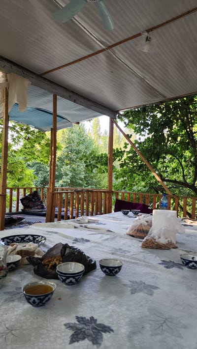 The image depicts an outdoor patio area, likely in Vodil, Uzbekistan, furnished with a table set for a meal or tea. The table is covered with a patterned cloth and features several small, decorative bowls, some containing a dark liquid, presumably tea. There are also bags of snacks, possibly nuts, and a large patterned plate. In the background, a wooden railing overlooks a lush, green natural setting with trees. A ceiling fan is visible overhead, attached to the corrugated roof of the patio structure. The scene suggests a relaxed, informal gathering outdoors.