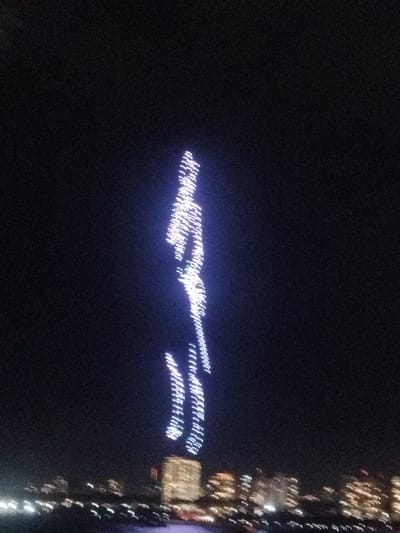 At night, a blurred cityscape sits at the bottom of the frame, topped by a dark sky where a vertical, luminous, elongated human-like shape is formed by clustered lights. The light formation is primarily white with a slight blue hue. The shape has distinct segments which might indicate the position of the limbs of the figure, though the overall impression is abstract due to blur.