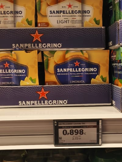 A display of San Pellegrino Limonata 33cl cans on a shelf in a grocery store located at coordinates (latitude: 50.834, longitude: 4.38) in Etterbeek, Belgium. The cans are stacked in two rows, with a price tag at the bottom, which reads "San Pellegrino Limonata 33cl, Meilleur prix 0,898€ (x5=5,39€), Le plus 2,72 cl." The cans are yellow with a red star and the San Pellegrino logo in blue. The text on the cans is in French and Italian, and it reads "Sanpellegrino Boissons pétillantes italiennes Limonata, Dal 1932." The shelf is white, and the background is a blue wall with the San Pellegrino logo in white. The cans are arranged in a neat and orderly fashion. There is no one visible in the image. The environment is clean and well-lit. The display is part of the beverage aisle in the grocery store. The price tag indicates a sale on San Pellegrino Limonata. The image is a still shot and there is no action taking place.