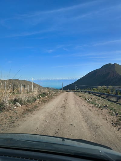 The image depicts a dirt road leading towards a mountainous area in Cholpon Ata, Kyrgyzstan. A blue sky with thin clouds is overhead. On the left side of the road, there's a fenced area with dry grass and rocks. In the distance, snowy mountain peaks and a body of water are visible. On the right side of the road, there is a green mountain slope, a large black metal pipe following the road edge, and some scattered trees. The image is taken from inside a vehicle, with a portion of the windshield visible at the bottom.