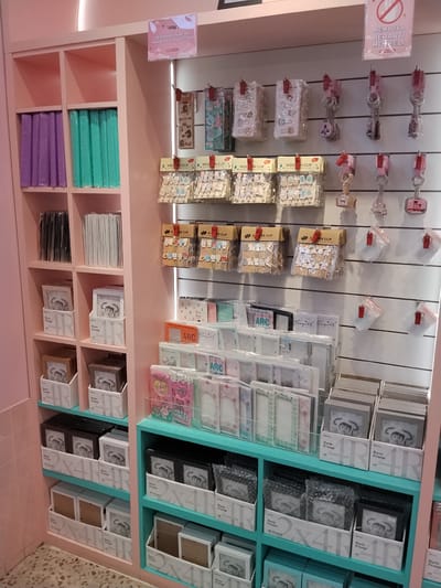 The image shows a retail display of stationery and small gift items in a store. The store appears to have pink shelving units and a turquoise display shelf. On the shelves, there are items such as photo frames, paper clips, keychains, and notebooks. Some of the items have text in what appears to be Indonesian. A sign at the top of the display reads "MEMBUKA BERARTI MEMBELI" which translates to "Opening means buying". The store is located in the city of Serang, Indonesia. There are no people visible in the image. The lighting is bright, suggesting it is daytime or the store is well-lit.