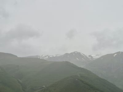 A wide-angle, eye-level shot captures a panoramic view of the majestic Caucasus Mountains in Stepantsminda, Georgia. The sky is overcast with soft, diffused light, creating a muted atmosphere. In the foreground, rolling green hills dominate the landscape, their slopes covered in lush vegetation. A narrow, dark ravine carves its way through the middle ground, adding a sense of depth. In the distance, rugged mountain peaks rise, some capped with patches of snow, indicating a high altitude. The scene appears peaceful and serene, with no visible signs of human activity except for a few small, indistinct structures nestled in the lower right corner of the image, suggesting a remote dwelling or settlement. The overall impression is one of natural beauty and tranquility, characteristic of a mountainous region.