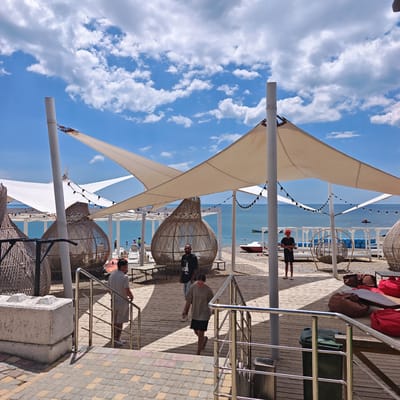 The image depicts a sun-drenched beachside seating area with a backdrop of the Black Sea. Several large, woven pod-like chairs are prominent in the foreground, offering shaded seating under expansive white canopies. String lights are suspended between poles, suggesting a venue that might operate into the evening. The ground is paved with wooden planks and decorative stone tiles. Several individuals are visible, some walking and others seated, interacting with the environment. In the distance, lounge chairs are arranged on the sand, and a few small boats are visible on the water. The sky is a vibrant blue with scattered white clouds. This scene is characteristic of beach resorts and cafes found along the coast of Sudak, Ukraine.