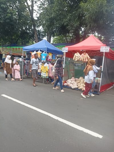 The image shows a street market in Purwokerto, Indonesia, located at the coordinates -7.418, 109.249. Two market stalls are set up on the side of the road, with one stall selling clothing and the other selling bags of fried snacks. The clothing stall has a blue tent and displays children's clothes on a rack. The snack stall has a red tent and is displaying bags of fried snacks on a rack. The road is paved with asphalt, and there is a white line painted down the middle of the road. Several people are walking past the stalls. One woman is wearing a pink dress, one woman is wearing a hijab, and one man is wearing a blue shirt and black pants. The market appears to be active with customers browsing the stalls. The scene is captured on a cloudy day.