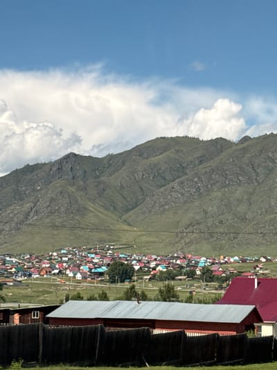 This image depicts a rural village nestled at the foot of a vast mountain range. The village features numerous small houses with brightly colored roofs in shades of red, blue, green, and white, creating a vibrant mosaic against the green landscape. A road winds through the village, with a few vehicles visible. In the foreground, a dark wooden fence runs across the bottom of the frame, partially obscuring two buildings. One building on the left is made of dark wood with a small window, and the other on the right has a red metal roof. The mountains in the background are covered in sparse vegetation, with rocky outcrops and deep ravines. The sky is a clear blue with scattered white clouds. The scene suggests a peaceful, remote settlement in a mountainous region. This scene is likely located in or near the city of Onguday, Russia, a region known for its mountainous terrain and traditional villages.