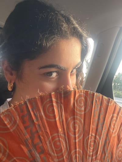 A close-up, eye-level medium shot shows a young person holding an orange fan in front of their face while sitting inside a vehicle. The person's dark, curly hair frames their face, with strands illuminated by sunlight. Their visible left eye, brown in color, is looking directly at the camera. A gold hoop earring is worn on the left ear. The fan, made of patterned orange fabric with lighter circular and geometric designs, obscures the lower half of their face. The background is blurred, showing the interior of a car with a gray headliner and a light-colored pillar with the word "Airbag" embossed on it. The scene appears to be set in Felgueiras, Portugal, with the exterior visible through a side window, hinting at a sunny day with trees and a road.