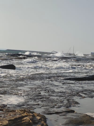This is a photograph of a rocky coastline with choppy water. The sky is overcast and pale. In the distance, there are construction cranes and buildings, suggesting an industrial or port area. Waves are crashing against the rocks, creating white foam. The foreground consists of wet, dark rocks with some shallow pools of water reflecting the sky. The overall impression is one of a somewhat industrial and weathered coastal environment. The location is specified as the city of Saiyn Şapağatov, Kazakhstan. There are no people or animals visible in the image.
