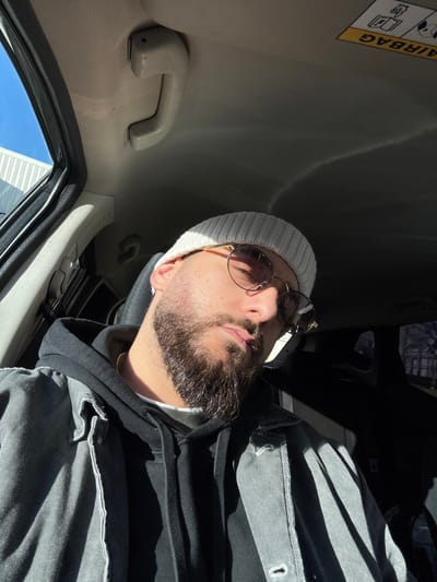 An adult male is positioned inside a vehicle, located in Hérouville-Saint-Clair, France. The subject is seated, wearing a light-colored knit beanie, round-framed sunglasses with tinted lenses, a black hooded sweatshirt, and a dark grey or faded black denim jacket. He has a full beard and mustache. Strong, direct natural light enters the vehicle from the left, illuminating the right side of his face (viewer's left) and casting the left side (viewer's right) into shadow. The interior features a beige headliner, a beige grab handle above the driver's side window, and an inverted "AIRBAG" warning label on the passenger's sun visor. Through the driver's side window on the left, a clear blue sky and a portion of a light-colored building with a corrugated facade are visible. The background visible through the passenger's side window on the right is darker and less defined, showing indistinct urban elements.