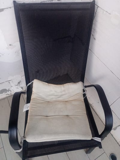 The image shows a close-up, eye-level view of an outdoor patio chair with a light-colored cushion. The chair has a black mesh back and seat with black metal armrests and legs. The cushion is centered on the seat and appears somewhat worn and creased. The background consists of a white, textured wall, likely made of concrete blocks, with some areas of exposed concrete or plaster. The floor beneath the chair is tiled with light-colored square tiles. The overall scene suggests an outdoor or semi-outdoor area, possibly a patio or balcony. The location is specified as Sovkhozny, Russia, though there are no visual cues within the image to confirm this specific location. There are no people or animals visible in the image.