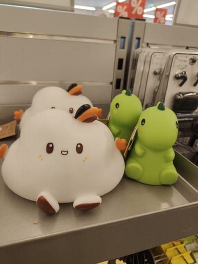 This is a close-up, eye-level shot of a retail display in a store, possibly a discount store, in Nowy Dwór Mazowiecki, Poland. The focus is on several novelty lamps in the foreground. On the left are two white, cloud-shaped lamps with simple cartoon faces, each with two brown eyes and a smiling mouth, adorned with orange and black duck-like accessories. To the right are two bright green dinosaur-shaped lamps, also with cartoon eyes. The items are arranged on a gray shelf. In the background, out of focus, are rows of merchandise in clear plastic packaging and red sale signs with "%" symbols. The lighting is bright and slightly diffused, typical of a retail environment.