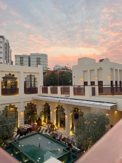 This image depicts an illuminated open-air courtyard at dusk, located within the Al Bait Sharjah hotel complex in Sharjah, United Arab Emirates. The view is from an elevated vantage point, looking down into the enclosed space and outwards towards the city skyline.
The courtyard is surrounded by cream-colored buildings featuring traditional Middle Eastern architectural elements, including arched window frames and decorative wooden latticework railings on upper levels and balconies. Multiple wall-mounted lanterns emit a warm, artificial light, contrasting with the natural light of the fading day.
A central, rectangular water feature, tiled in a dark teal, occupies the lower part of the courtyard and is equipped with internal lights. Surrounding this feature are various outdoor seating arrangements: a larger dining table with wicker chairs and a sectional sofa with red cushions to the left, and two smaller round tables each with two ornate chairs to the right. Small trees and potted plants are integrated into the landscaping around the seating areas. The ground features a geometric tile pattern.
One person is observed seated on an upper terrace, partially obscured by a wooden pergola. No other individuals are distinctly visible within the courtyard's main seating areas.
The background reveals contemporary multi-story buildings against a twilight sky. The sky is characterized by an altocumulus cloud formation, illuminated with shades of pink, orange, and light blue, consistent with a sunset.
