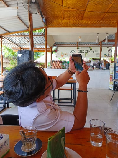 A person, viewed from behind, is seated at a wooden table, holding up a smartphone to take a picture. The person wears a light-colored collared shirt and glasses. On the table, there are two glasses, one partially filled with water, a coaster with a small glass, a packet of tissues, and what appears to be food wrapped in banana leaves. In the background, there is a counter area with coffee-making equipment, signage that reads "EDK Kuphi", and stacks of cans. The setting appears to be an outdoor cafe or restaurant with a bamboo-thatched roof and ceiling fans. The location is identified as Julikeude Dua, Indonesia.