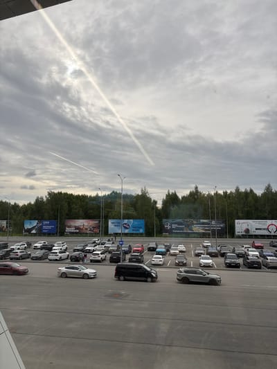 This image shows a parking lot filled with cars, viewed from an elevated angle. The cars are parked in rows, suggesting a public space like a shopping center or event venue. In the background, there is a line of trees and a cloudy sky with a distinct contrail from an aircraft. Several billboards are visible along the edge of the parking lot, featuring Russian text related to various businesses and services, including "Союз" (Union), "Безопасность на высоте" (Safety at height), "Чистое небо" (Clear sky), and "Чистая вода" (Clean water). The overall scene appears to be a daytime setting, with the lighting suggesting it might be overcast or late afternoon. The presence of multiple cars and billboards indicates a commercial or public area.