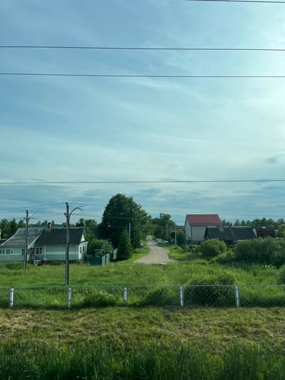 The image depicts a rural scene in Chudovo, Russia, viewed from a train window. The perspective is a low-angle shot, with the image rotated 90 degrees counterclockwise, giving the impression of looking out horizontally from a moving vehicle. The foreground is dominated by the sky, which is a pale blue with scattered white clouds, suggesting daytime. Three dark, thin electrical wires stretch diagonally across the sky, indicating the presence of power lines.
To the right of the sky, a grassy field stretches along a chain-link fence. Beyond the fence, there is a residential area with several houses. The houses are predominantly light green with white trim and dark roofs. One house in the mid-ground has a distinctive red roof. There is a dirt road winding through the area, and a silver car is parked on it. Trees are scattered throughout the residential area and along the edge of the field, contributing to a lush, green landscape.
The overall impression is one of a tranquil, sunny day in the Russian countryside. The focus is on the natural landscape and the simple architecture of the rural dwellings. No people are clearly visible, and there are no discernible emotions conveyed. The time of day appears to be late morning or early afternoon, given the brightness of the sky. The location cues, such as the style of the houses and the general landscape, are consistent with a rural Russian setting. There is no visible text in the image.