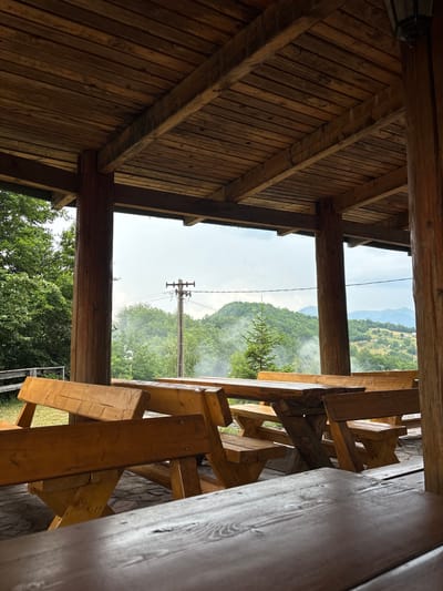 The image depicts an outdoor seating area constructed of wooden picnic tables and benches, positioned under a rustic wooden roof supported by thick, rough-hewn wooden posts. The tables and benches are made of natural wood, some with a light orange-brown stain, and others with a darker, more weathered finish. The flooring appears to be cobblestone or irregularly shaped stones.
Beyond the seating area, the view opens up to a lush, green landscape of rolling hills and mountains, densely covered with trees. In the middle ground, a single wooden utility pole with wires is visible, and wisps of smoke or mist are present in the air, adding a hazy quality to the distant scenery. The sky is overcast, suggesting a cloudy or possibly rainy day. The overall impression is of a secluded, natural setting, likely a rural picnic spot or an outdoor cafe in a mountainous region. The location is specified as Mekića Kula, Montenegro. There are no people or animals visible in the image.
