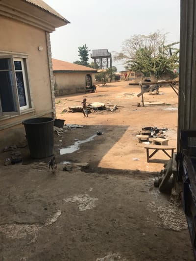 The image depicts an unpaved outdoor communal area in Oba-Ile, Nigeria, under a hazy daylight sky. In the immediate foreground left, the side of a light-colored building with a light brown tiled roof is visible, featuring a partially open white-framed window. Adjacent to this building, a large, dark grey fluted plastic refuse bin stands on a concrete patch. A small, multi-colored chicken walks on the concrete near the bin.
The ground transitions from concrete to packed dirt/sand, marked by several puddles and damp streaks. A young child, barefoot and wearing light-colored shorts, walks leftward across the dirt mid-ground, holding a small, light-colored bowl or dish in their left hand. Around the child and extending into the mid-ground, scattered debris, including wooden planks, various scraps, and small containers, are present.
Further into the mid-ground, to the left, another single-story building with a darker, possibly green corrugated metal roof and a dark, ornate metal gate is visible. Near this building, a person, dark-skinned, is partially visible in a bent-over posture. Towards the right mid-ground, a low, rough wooden bench and other makeshift wooden structures are situated amongst additional debris.
In the background, a tall metal water tower with a tank is prominent, positioned to the left of a large, sparsely leafed tree. Other distant low-rise buildings are visible, with faint figures potentially present near them. The overall impression is of a lived-in, utilitarian space with elements of both established structures and informal arrangements.