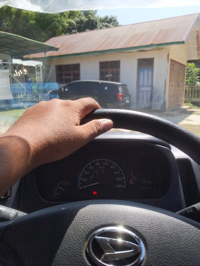 In Gampongpande, Indonesia, a hand is resting on the steering wheel of a vehicle. The interior of the vehicle is visible, including the dashboard with a speedometer. Outside the vehicle, a building with a corrugated metal roof and white walls is seen, along with a dark-colored car parked in front of it. Sunlight illuminates the scene, casting shadows.