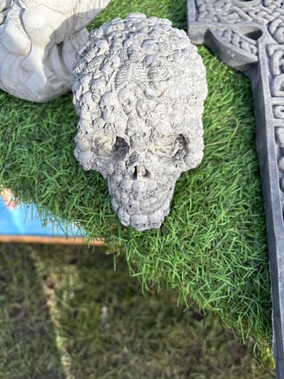 A high-angle shot shows an ornamental skull, sculpted from a gray material, seemingly stone, placed on artificial green grass. The skull's surface is textured with small skull-like shapes, indicating a possible theme of mortality. Adjacent to the skull is a dark gray decorative architectural element, possibly part of a larger structure. Beneath the artificial grass, a patch of blue and brown is visible.
Without additional context, it's impossible to confirm the location specifically in Chirk, United Kingdom. However, the image seems to depict a garden or decorative setting.