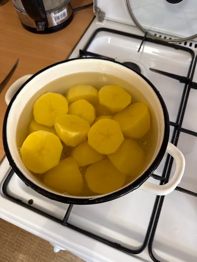 A white enamel pot filled with water and sliced potatoes sits on a white gas stove. The potatoes are cut into thick, irregular rounds and are submerged in the clear liquid. The pot has a black rim and two white handles. The stove has four black grates. A partial view of a silver electric kettle and a wooden countertop is visible in the upper left corner. The scene appears to be a domestic kitchen setting. The location is specified as Морозовка, Russia.