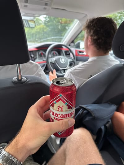 The image depicts the interior of a Volkswagen vehicle, located in Catwick, United Kingdom. The perspective is from the rear passenger compartment, looking forward. In the foreground, a right hand, wearing a silver-colored wristband or watch, holds an opened red Cruzcampo beer can with its pull tab elevated. The can displays a red and white geometric pattern and the brand name "Cruzcampo SEVILLA". Below the can, the individual's bare legs are partially visible.

In the driver's seat, a person with short, curly brown hair and wearing a white shirt is visible from behind, with their right hand on the steering wheel. The car's dashboard features red trim accents, and the steering wheel bears a Volkswagen emblem. The instrument cluster is partially visible with red needles. A second individual, also in a white shirt, occupies the front passenger seat, their torso partially visible. Through the vehicle's windshield, blurred green foliage is discernible, indicating an outdoor, natural or suburban environment during daylight hours. A dark blue bag is visible on the floor or seat in the lower right quadrant of the image.