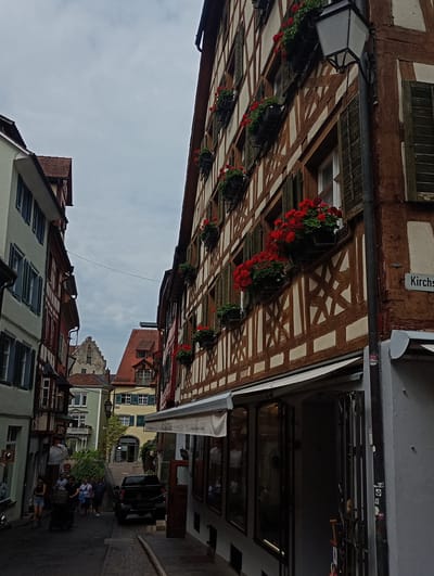 This image captures a picturesque street scene in Meersburg, Germany, with traditional half-timbered buildings lining the narrow cobblestone lane. The architecture is characterized by exposed wooden beams against a lighter plaster or whitewashed background, a common feature of historic German towns. Red geraniums spill abundantly from window boxes on multiple levels of the buildings, adding vibrant splashes of color against the rustic facades.
In the foreground, a dark-colored SUV is parked on the right side of the street, partially obscuring the entrance to a shop with large glass windows and an awning. On the left side, several people are walking down the street, some with shopping bags, suggesting a commercial area or a popular tourist destination. A person is pushing a stroller, and another individual is seen walking a dog. The street itself is paved with cobblestones, indicative of older European cities.
Further down the street, more buildings are visible, including one with a green facade and a balcony adorned with greenery. In the distance, a more imposing stone building with a stepped gable roof rises above the surrounding structures, possibly a historical landmark or part of the town's castle complex. The sky is overcast, casting a soft, diffused light on the scene, which contributes to a slightly muted but charming atmosphere. A street sign reading "Kirch..." is affixed to the wall of a building on the right, hinting at the street's name. The overall impression is one of a quaint and well-preserved historic European town.