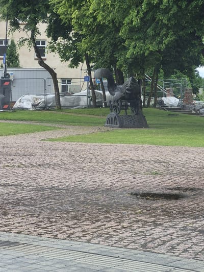 The image captures an outdoor scene in Šilutė, Lithuania, featuring a park-like area with a cobblestone path in the foreground and a sculpture in the middle ground. The sculpture appears to be an animal, possibly a mythical creature, mounted on a wheeled base, made of dark metal. Behind the sculpture, there are trees with dense green foliage, and in the background, a building with windows and a white temporary structure covered in sheeting. Construction fencing and materials are also visible, suggesting ongoing work in the area. The cobblestone path is wet in places, indicating recent rain. The lighting suggests an overcast day.