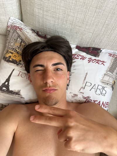 A young man with dark hair and a Jordan headband lies on a pillow with a New York and Paris pattern. He is looking at the camera and winking, with his right hand held up, displaying a peace sign. The background appears to be a sofa. The setting is indoors. The location of Marikostinovo, Bulgaria, is not depicted in this image.