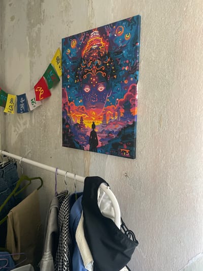 An indoor scene shows a wall with a painting and prayer flags. The painting depicts a cosmic landscape with a large, stylized face. Below the painting, a clothing rack holds various garments. To the left of the painting, a string of colorful prayer flags hangs against the textured wall. The wall appears to be made of concrete with peeling paint. The overall environment suggests a domestic setting. The location of Kopeysk, Russia is not determinable from the visual evidence.