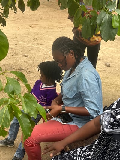 A medium shot captures three individuals seated on a wooden bench in Ganyingbo, Nigeria. In the foreground, a young child wearing a purple sweater with pink trim that reads "KESNA SCHOOLS" sits to the left, looking to the right. To the child's right, an adult woman in a light blue denim shirt and red pants is looking down at a smartphone in her hands. To the far right, another person is partially visible, dressed in a black and white patterned outfit. The ground is sandy with scattered small stones and sparse vegetation. Overhead, large green leaves from a tree obscure parts of the background. The scene appears to be outdoors, possibly in a schoolyard or a casual gathering area.