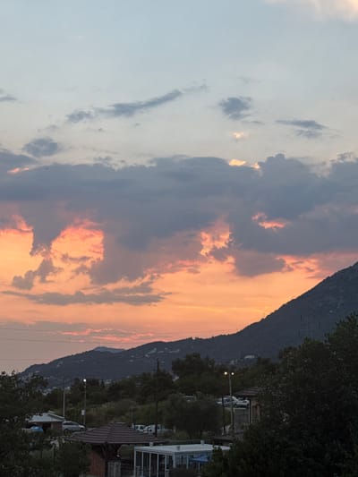 The image depicts a sunset scene in Sustaš, Montenegro. The sky is filled with dramatic clouds, painted in hues of orange, pink, and gray, suggesting the end of the day. A prominent, dark mountain range dominates the right side of the frame, its silhouette outlined against the vibrant sky. 

Below the mountain, a line of trees runs along the landscape, transitioning into a residential area. Several houses with dark tiled roofs are visible, along with parked vehicles and streetlights. The presence of power lines and a distant transmission tower on the mountain further indicate a populated area.

The overall mood is serene and picturesque, capturing the natural beauty of the sunset juxtaposed with the man-made structures of the town. The time of day is clearly evening, given the warm, fading light. The weather appears calm, with no indication of rain or strong winds. There is no visible text in the image.